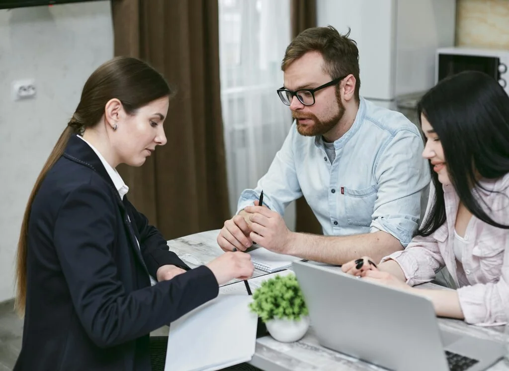 Three people at a table discuss documents. A woman in a blazer points to a paper, while a man with glasses and another woman look on intently, making an informed decision for Jubilee Life Insurance. A laptop and small potted plant sit on the table in the neutral-toned room with a window in the background.