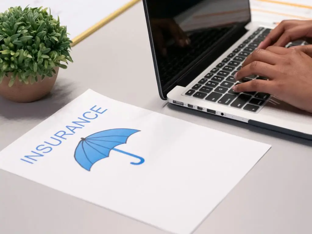 A person types on a laptop with a small potted plant beside it. Next to the laptop is a piece of paper featuring a blue umbrella graphic and the words "The Top 5 Insurance Companies" printed on it.