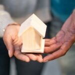 Close-up of two pairs of hands holding a small wooden house model. The hands of two individuals, one in a gray sweater and the other in a blue shirt, are gently supporting the light-colored miniature house together, symbolizing unity and trust—ideal for promoting the best home insurance companies in Pakistan.