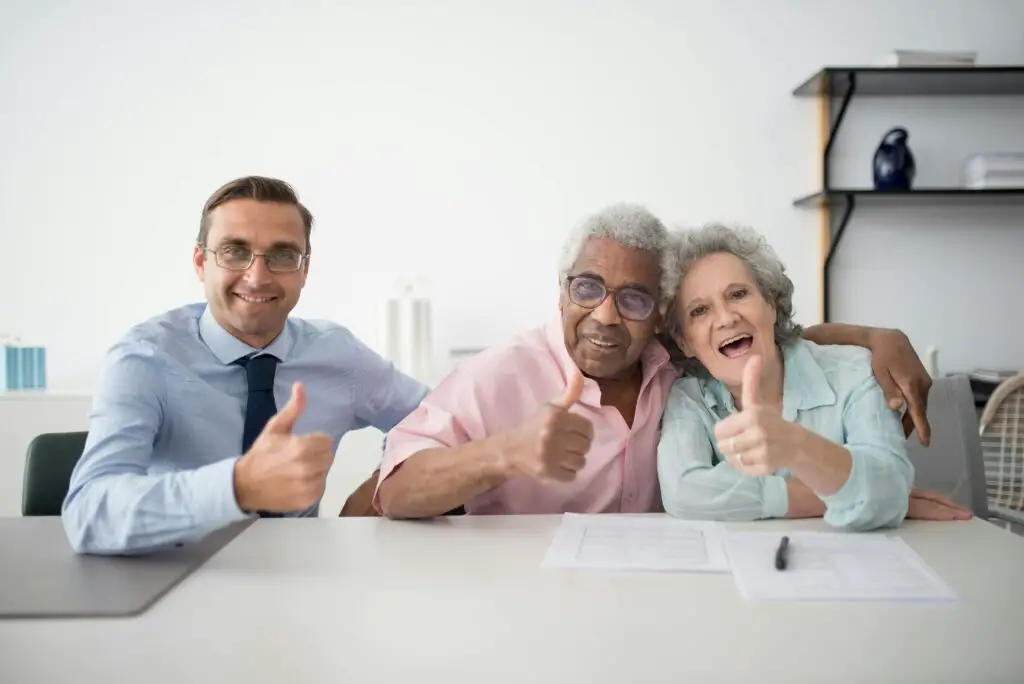 Three people are sitting at a table giving thumbs up. On the left, a man in glasses and a blue shirt smiles at the camera. In the middle, a senior man in a pink shirt and glasses, smiles while embracing a senior woman on the right who is also smiling and wearing a light blue shirt. Documents from Adamjee Insurance are on the table.