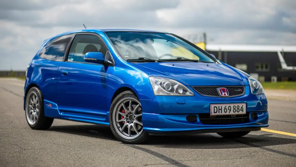 A blue Honda Civic Type R hatchback is parked on an open road under a cloudy sky. The car features alloy wheels and aerodynamic design elements, with the license plate "DH 69 884." A building is visible in the distance, reminding you to ensure your auto insurance is always up to date.