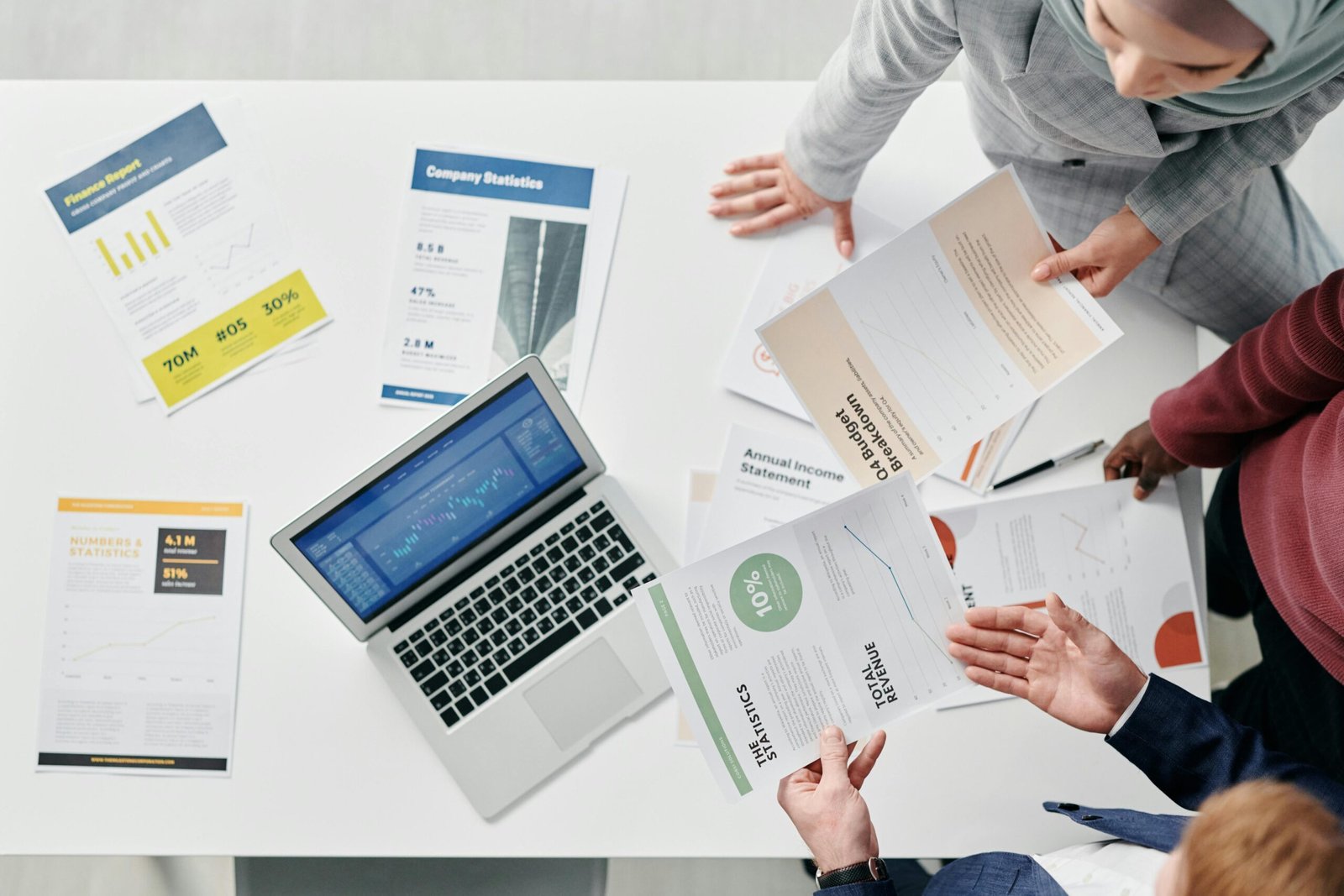 A group of people in business attire are gathered around a table, reviewing various financial documents and charts. A laptop displaying data is open on the table, along with printed reports and graphs. The scene depicts a collaborative business meeting.