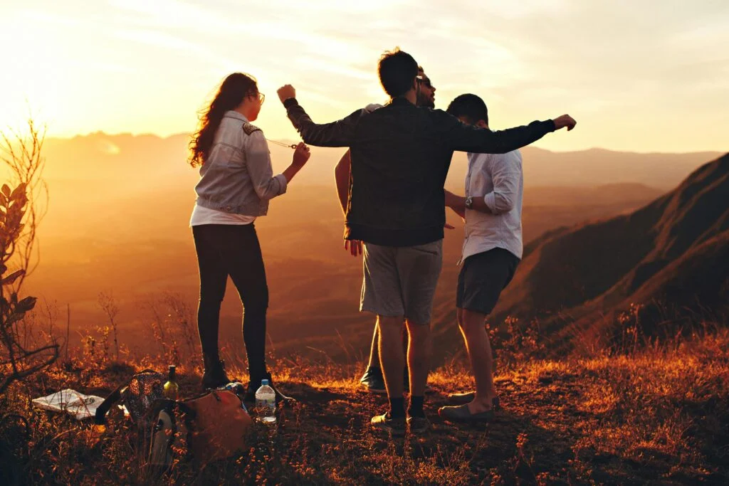 Four people are standing on a hilltop during a beautiful sunset, with two of them embracing while the others are nearby. They appear to be enjoying the view. In the foreground, there are some bags and bottles on the ground, as if they paused to cherish life's golden moments—just like good life insurance can do.