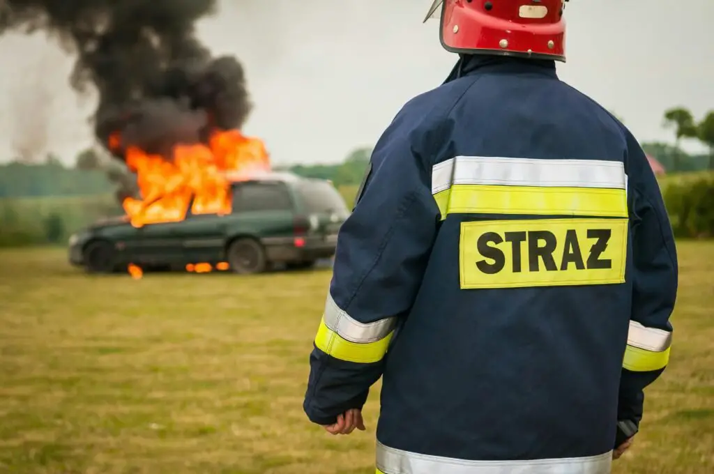 A firefighter wearing a red helmet and a dark blue jacket with the word "STRAŻ" in yellow stands on grass, facing a burning vehicle in the background. Thick black smoke rises from the engulfed car, highlighting the importance of having reliable Auto Insurance like IGI Auto Insurance to cover unexpected events.