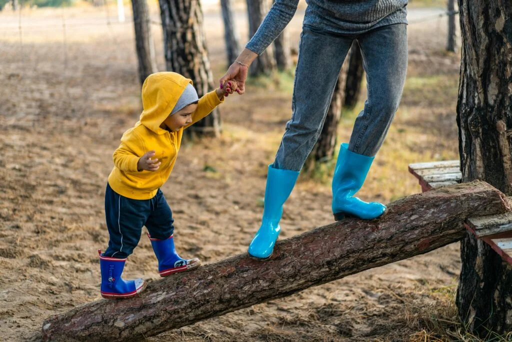 A toddler wearing a yellow hoodie and blue rain boots is balancing on a fallen tree trunk, holding an adult's hand for support. The adult is wearing blue rain boots and standing next to the child, guiding them through a wooded area.