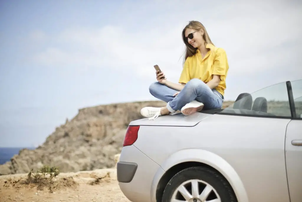 A woman wearing sunglasses, a yellow shirt, and jeans sits cross-legged on the trunk of a silver convertible car. She is smiling and looking at her phone, perhaps checking out the best insurance company. The background features a rocky cliff and a clear blue sky.