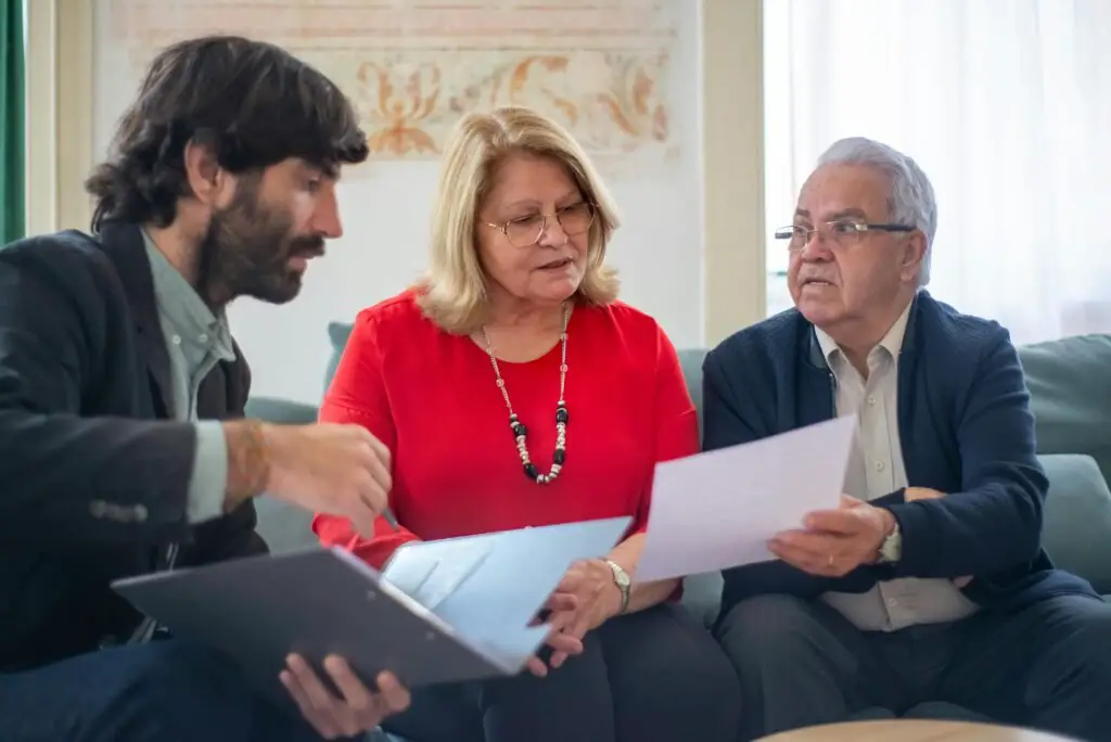 Three people are sitting and engaged in a discussion about home insurance in Pakistan. The person on the left holds a laptop, the person in the middle is holding a document, and the person on the right looks on attentively. They appear to be in an indoor setting with a couch and bright background.