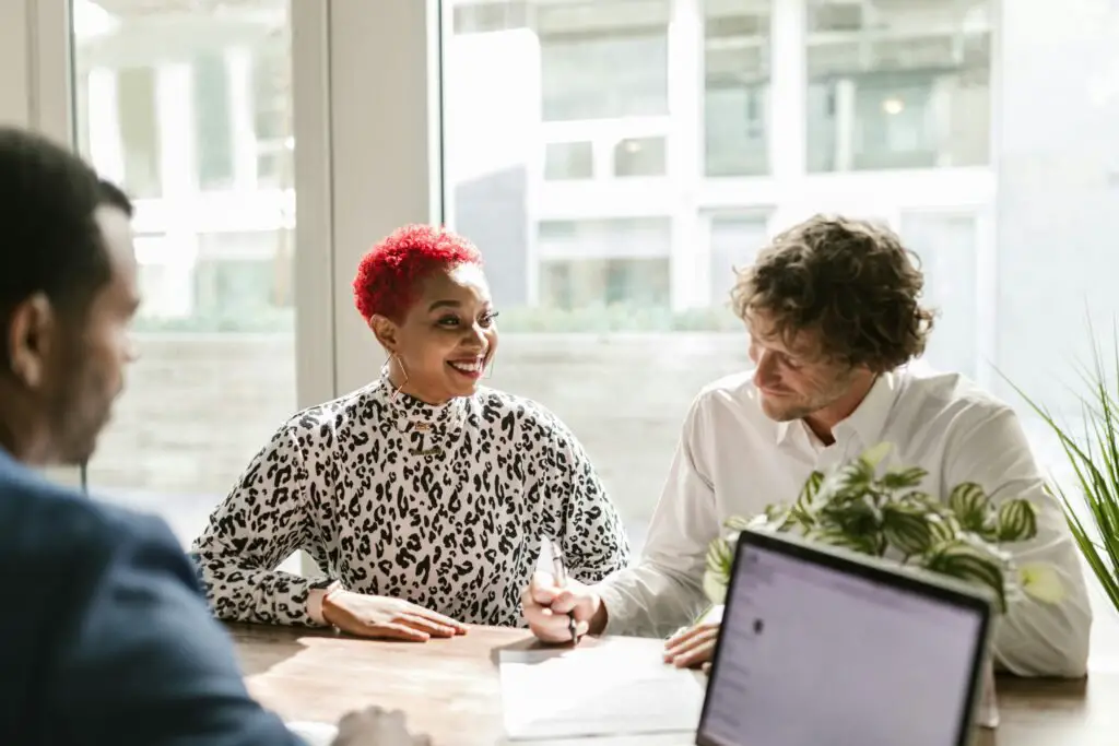 Three people sit around a desk engaged in a discussion. One person with bright red hair and a leopard print shirt smiles brightly, while the person beside them, wearing a white shirt, takes notes. A third person is partially visible, with a laptop open in the foreground showing EFU Life Insurance benefits as their guide.