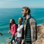 A group of four friends, laughing and talking, stand on a cliff overlooking a vast ocean under a clear blue sky. Everyone is dressed casually in outdoorsy clothes, clearly enjoying a sunny day of hiking or exploring. The sea stretches out to the horizon behind them.