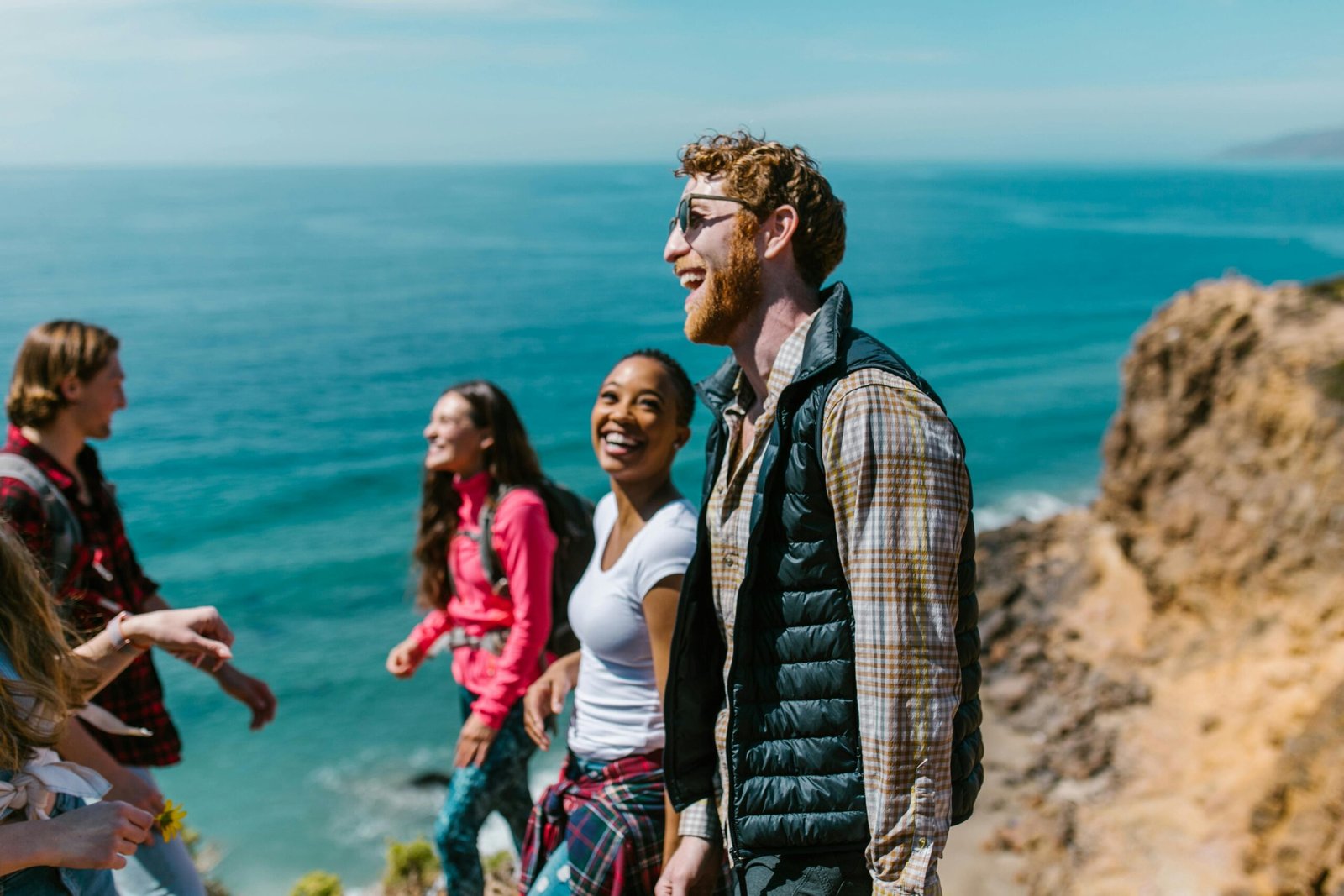 A group of four friends, laughing and talking, stand on a cliff overlooking a vast ocean under a clear blue sky. Everyone is dressed casually in outdoorsy clothes, clearly enjoying a sunny day of hiking or exploring. The sea stretches out to the horizon behind them.