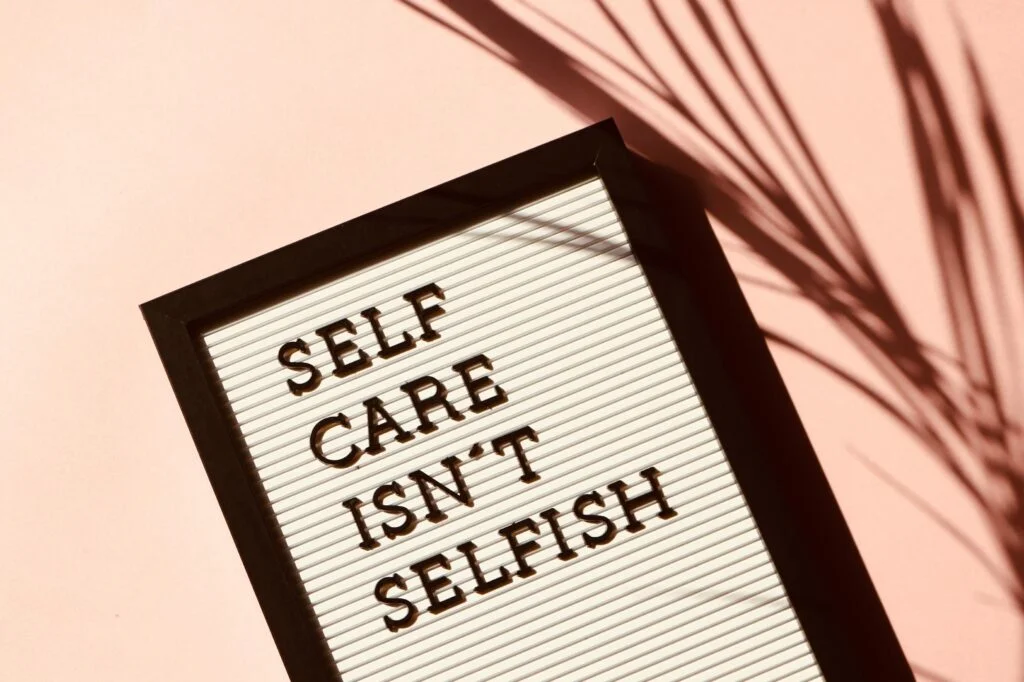 A felt letter board with black letters reads "SELF CARE ISN'T SELFISH" against a light pink background. The board has a black frame, and a shadow of a plant falls on the right side of the image, adding a natural touch.