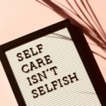 A felt letter board with black letters reads "SELF CARE ISN'T SELFISH" against a light pink background. The board has a black frame, and a shadow of a plant falls on the right side of the image, adding a natural touch.