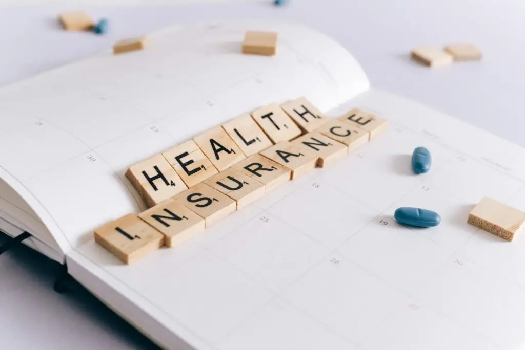 Scrabble tiles arranged to spell "HEALTH INSURANCE" are placed on an open planner calendar. A few light blue pills are scattered around the planner, reflecting the importance of OPD Health Insurance in managing daily health needs in Pakistan.