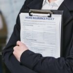 Person wearing a dark blazer, holding a clipboard with a "Home Insurance Policy" document. The policy includes sections for personal details and previous insurance history. The background is blurred, focusing attention on the clipboard detailing home insurance coverage.