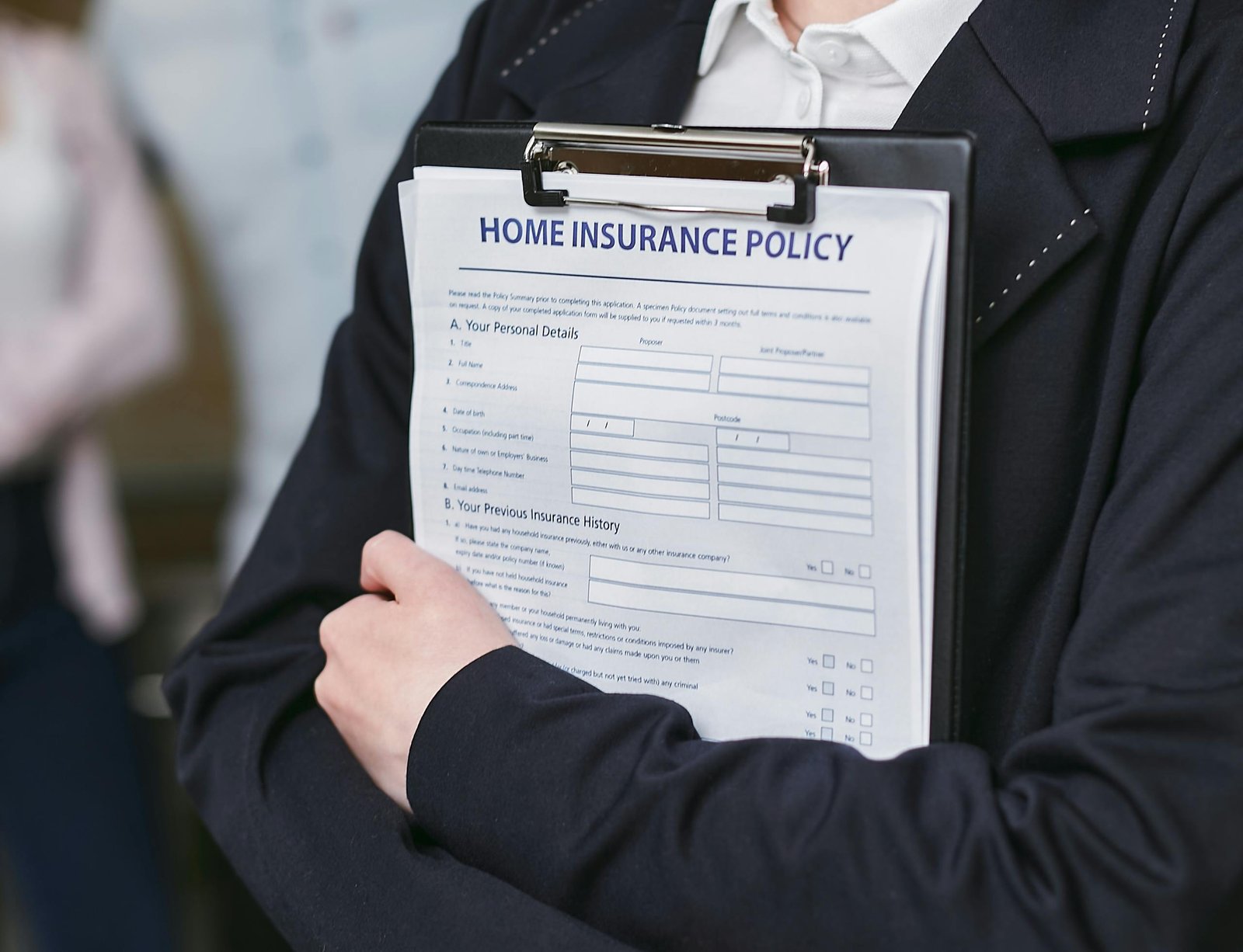 Person wearing a dark blazer, holding a clipboard with a "Home Insurance Policy" document. The policy includes sections for personal details and previous insurance history. The background is blurred, focusing attention on the clipboard detailing home insurance coverage.