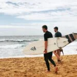 Two surfers in wetsuits carry their white surfboards as they walk toward the ocean on a sandy beach. Waves are crashing in the background, and a few people are in the water ahead. The sky is partly cloudy.