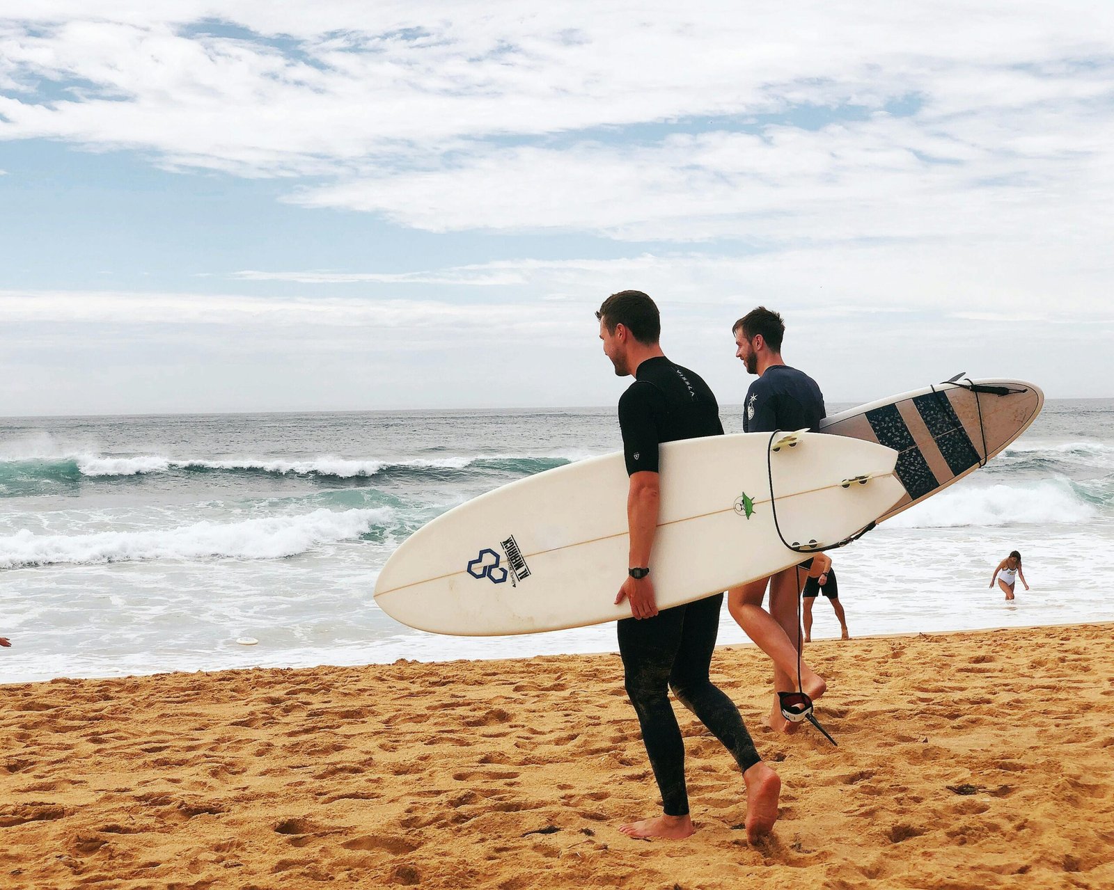 Two surfers in wetsuits carry their white surfboards as they walk toward the ocean on a sandy beach. Waves are crashing in the background, and a few people are in the water ahead. The sky is partly cloudy.