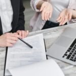 Two individuals are discussing a document on a table. One person, whose face is not visible, is pointing to the document with a pen. The other, also partially obscured, has their hands open in a questioning gesture. Beside the document and laptop, they delve into how to secure insurance for their business.