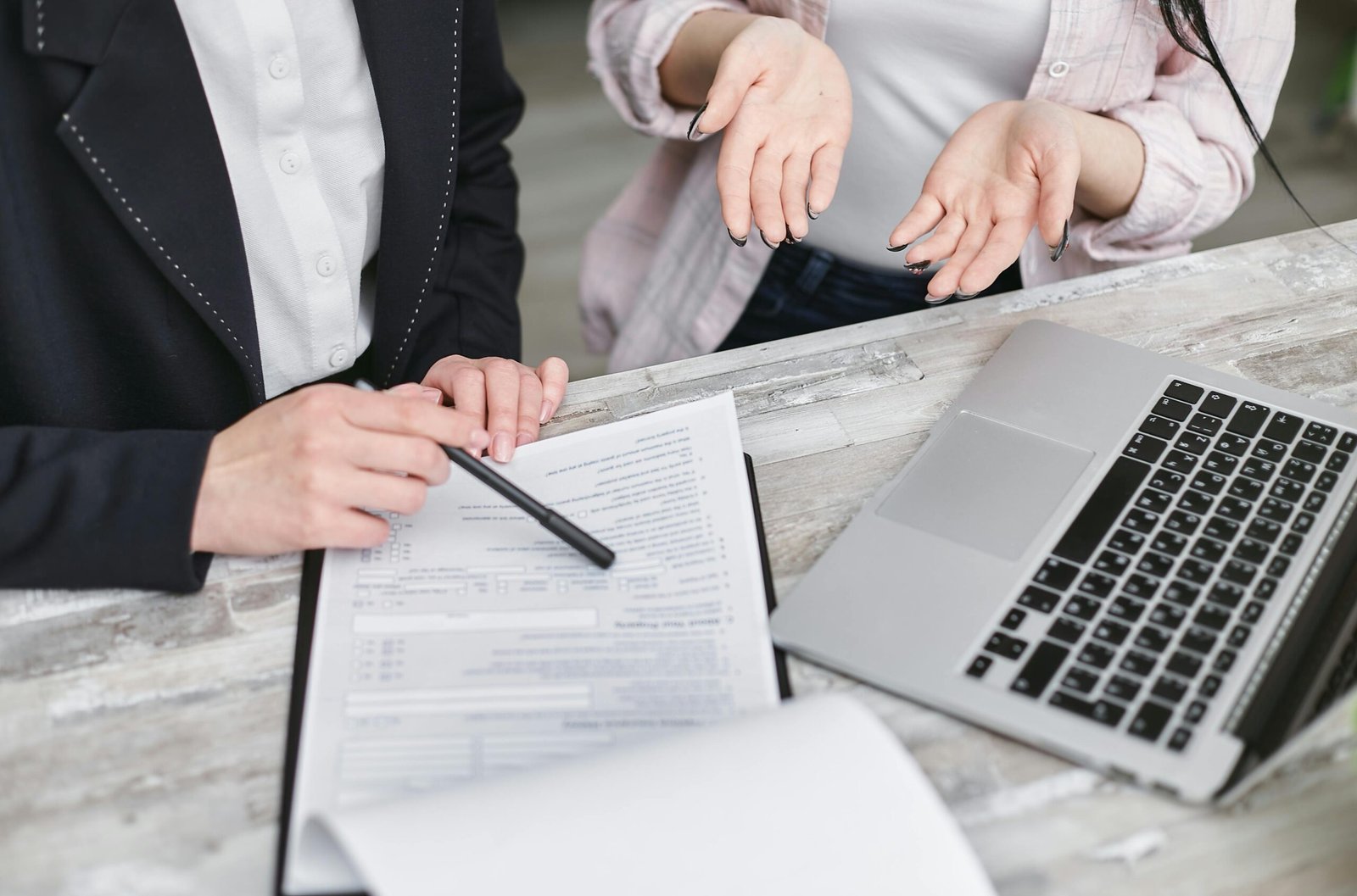 Two individuals are discussing a document on a table. One person, whose face is not visible, is pointing to the document with a pen. The other, also partially obscured, has their hands open in a questioning gesture. Beside the document and laptop, they delve into how to secure insurance for their business.