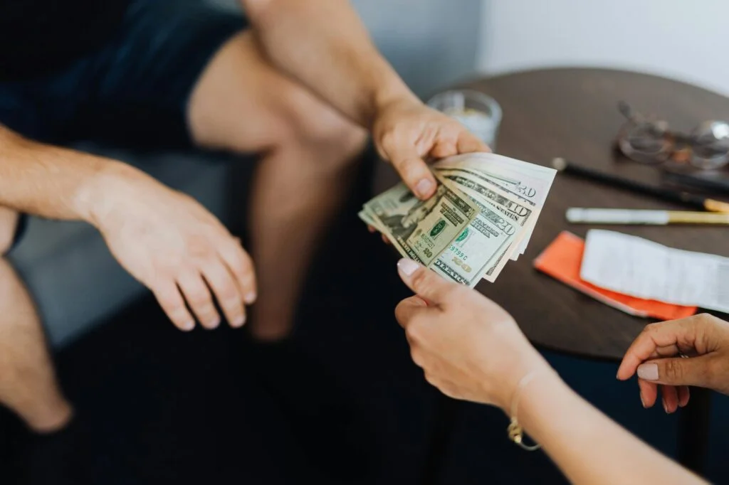 Two people exchanging a stack of U.S. dollar bills over a dark round table, which also holds a pair of glasses, a pen, a receipt, and an orange-wrapped object. Only their hands and part of their arms are visible. This scene might depict a transaction requiring small business insurance to ensure both parties are covered.