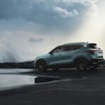 A teal SUV is parked on a rocky shore next to a calm body of water. The sky is filled with dramatic clouds, and waves crash against the rocks in the background.