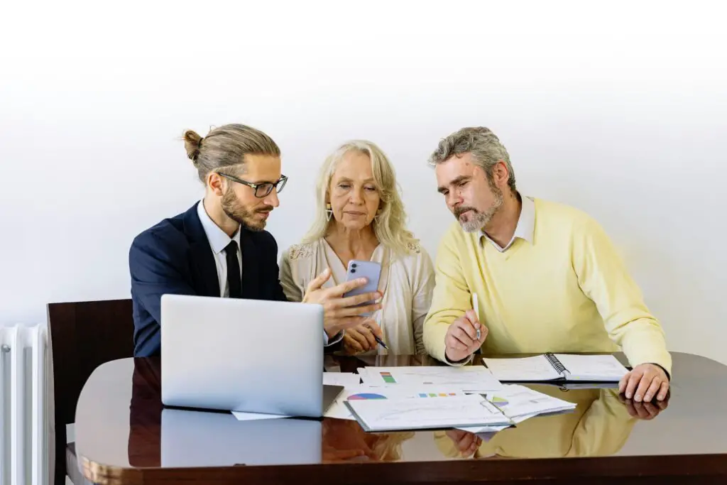 A businessman with a laptop, wearing a suit and glasses, is sitting at a table with an older couple. The woman is looking at a smartphone while the man holds a pen and examines documents spread out on the table. They seem to be discussing the benefits of third-party insurance in their meeting.