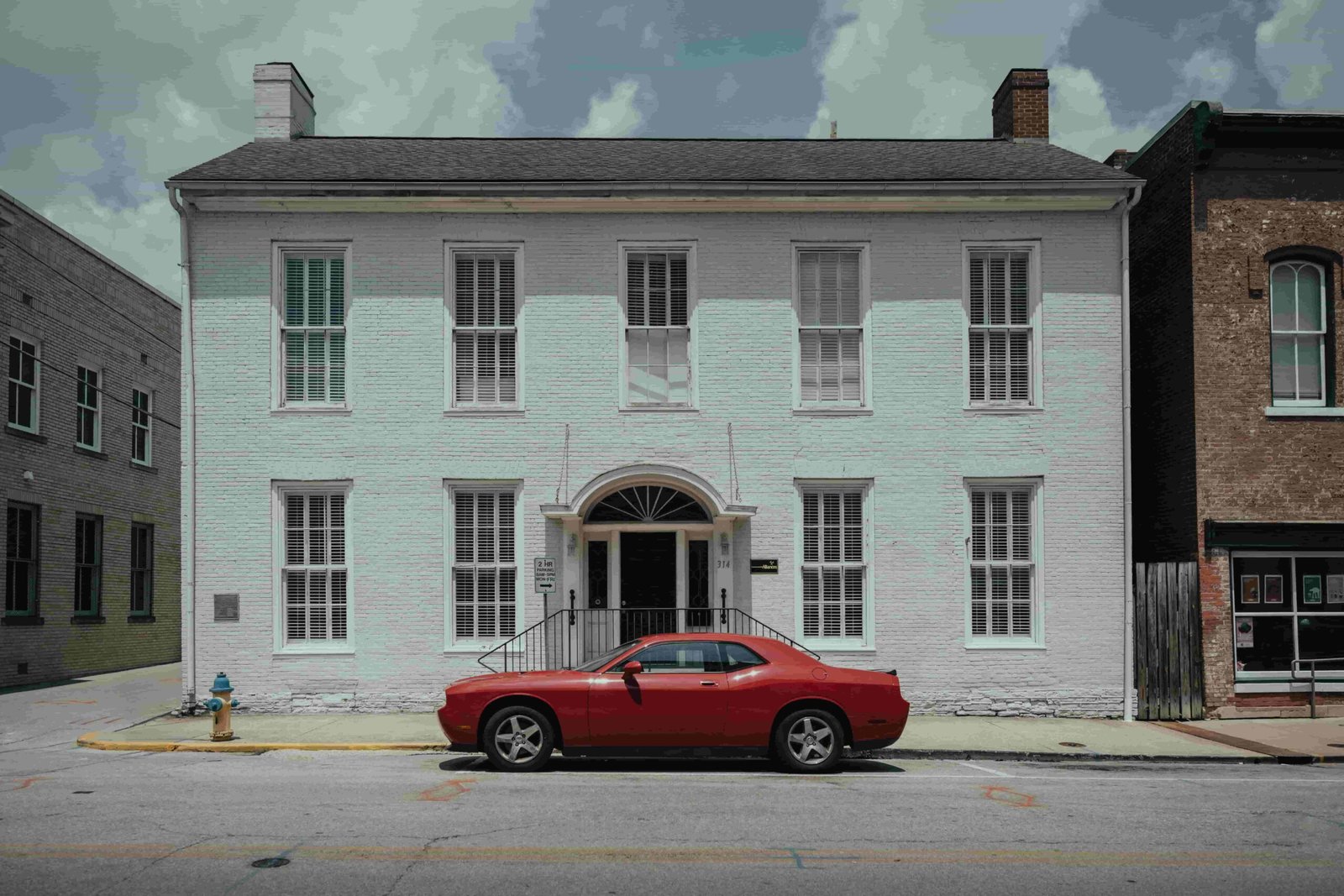 A red car is parked in front of a two-story white brick building with multiple windows. The building, possibly requiring insurance, has a central arched entrance with a black handrail leading up to it. The scene is set on a quiet street with neighboring buildings on either side.