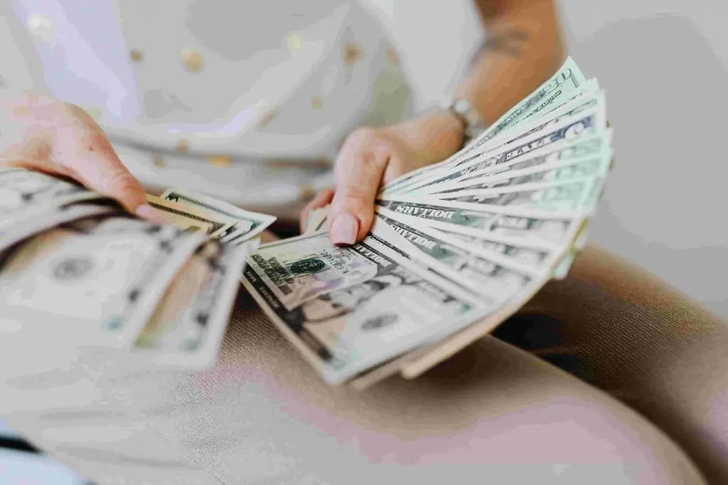 A person holding and fanning out a stack of U.S. dollar bills in their hands, possibly celebrating a successful insurance deal. The individual is wearing a white shirt and light-colored pants. The photo primarily focuses on the money and hands, with a blurred background.