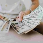 A person holding and fanning out a stack of U.S. dollar bills in their hands, possibly celebrating a successful insurance deal. The individual is wearing a white shirt and light-colored pants. The photo primarily focuses on the money and hands, with a blurred background.