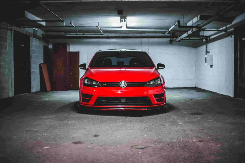 A red Volkswagen hatchback is parked in an empty, dimly-lit underground parking garage. The car's front is facing the camera, showcasing its headlights and grille. The concrete walls and ceiling pipes are visible, emphasizing the industrial setting that makes you appreciate the need for good insurance.