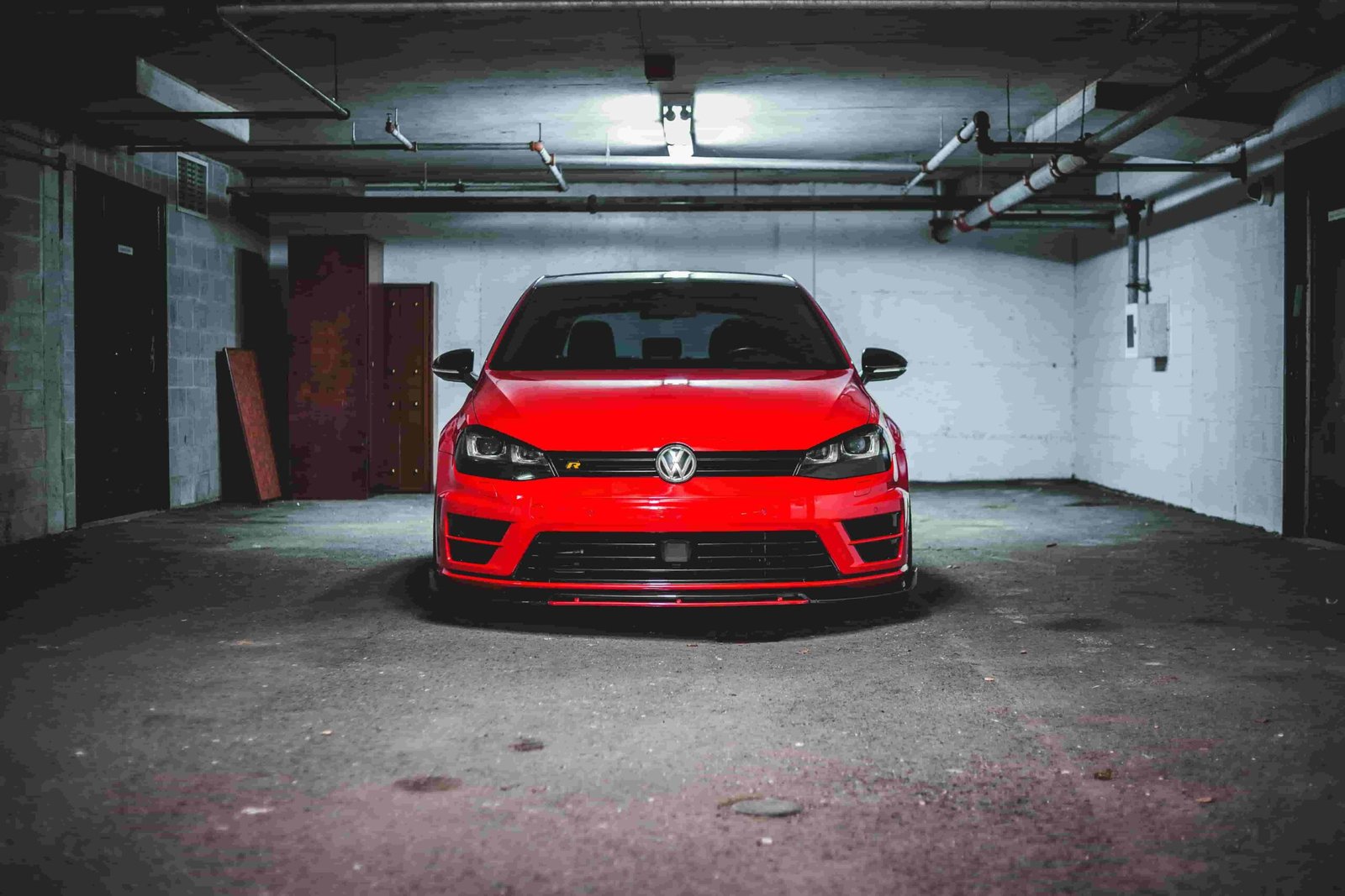 A red Volkswagen hatchback is parked in an empty, dimly-lit underground parking garage. The car's front is facing the camera, showcasing its headlights and grille. The concrete walls and ceiling pipes are visible, emphasizing the industrial setting that makes you appreciate the need for good insurance.