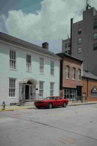 A red car, likely with insurance, is parked on the side of a street in front of a row of buildings, including a white two-story building and a brown brick building. The street is empty, and there are a few fluffy clouds in the sky.