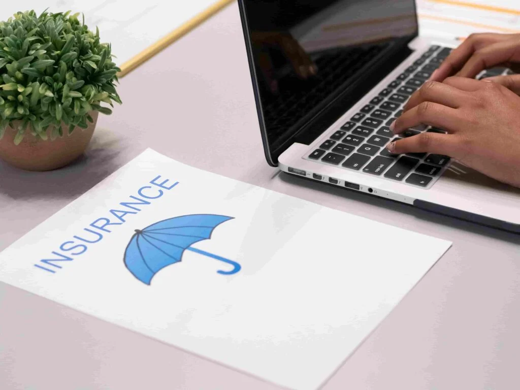 A person types on a laptop keyboard next to a paper with the word "INSURANCE" and an image of a blue umbrella on it. A green potted plant is in the background on a light-colored desk.