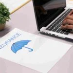 A person types on a laptop keyboard next to a paper with the word "INSURANCE" and an image of a blue umbrella on it. A green potted plant is in the background on a light-colored desk.