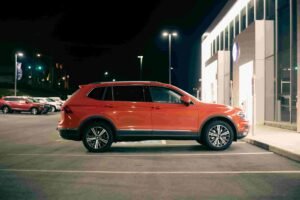 A red mid-size SUV is parked in a well-lit parking lot at night, its sleek frame standing out next to a building with large, illuminated windows. Other vehicles are visible in the background, each one likely covered by comprehensive insurance.