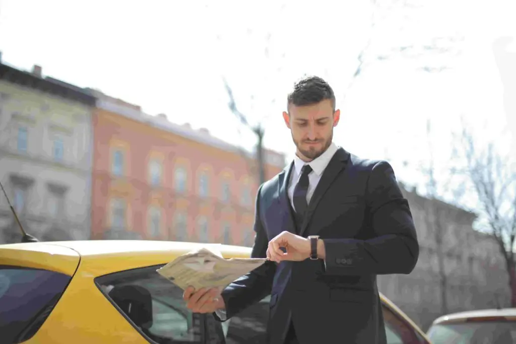 A man in a suit stands next to a yellow car on the street, holding several newspapers in one hand while checking his watch with the other. With a serious expression, he looks like he's heading to an important insurance meeting. Buildings with various facades are visible in the background.