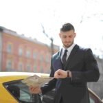 A man in a suit stands next to a yellow car on the street, holding several newspapers in one hand while checking his watch with the other. With a serious expression, he looks like he's heading to an important insurance meeting. Buildings with various facades are visible in the background.