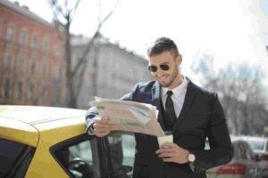 A man in a black suit and sunglasses stands outdoors beside a yellow car, smiling while reading a newspaper. He is holding a coffee cup in one hand and leaning casually against the car. With the confidence of someone who knows his insurance is rock-solid, he enjoys his morning read.