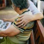 Two older adults sit on a bench, one reading a newspaper. The reader, with short gray hair and wearing a striped shirt, is lovingly embraced by the other, who shows a hand with a wedding ring. In the background, an insurance vehicle is parked.