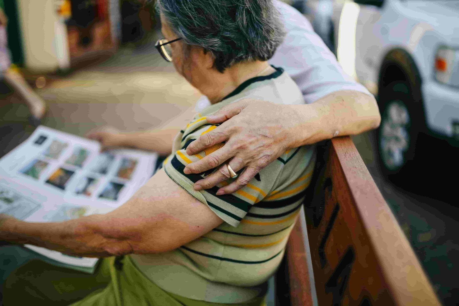 Two older adults sit on a bench, one reading a newspaper. The reader, with short gray hair and wearing a striped shirt, is lovingly embraced by the other, who shows a hand with a wedding ring. In the background, an insurance vehicle is parked.