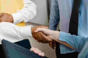 Two individuals, one in a white shirt and the other in a blue shirt, are shaking hands in a professional setting. Nearby, a third person in a light blue shirt and dark tie looks on, with part of an insurance document visible on the laptop in the foreground.