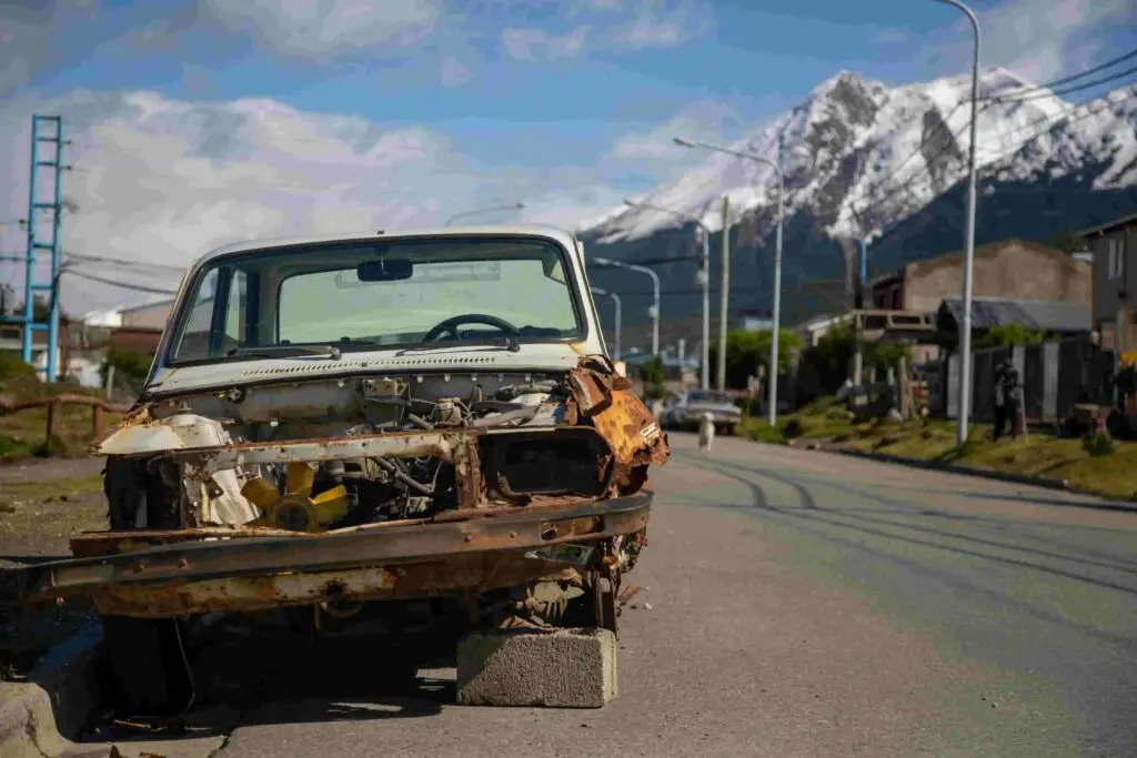 A rusted, dismantled car sits abandoned on blocks by the side of a road with a view of snow-capped mountains in the background. Missing its front grill and headlights, it stands as a poignant reminder of urban decay meeting natural beauty, a stark contrast to the pristine promise often seen in insurance commercials.