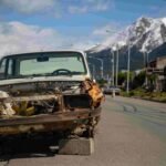 A rusted, dismantled car sits abandoned on blocks by the side of a road with a view of snow-capped mountains in the background. Missing its front grill and headlights, it stands as a poignant reminder of urban decay meeting natural beauty, a stark contrast to the pristine promise often seen in insurance commercials.