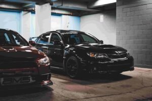 Two cars are parked in a dimly lit garage. On the left is a red car, partially visible, and to the right is a black car with insurance stickers on the windshield. The black car has a sporty appearance with a hood scoop and dark wheels, casting a strong reflection on the polished floor.