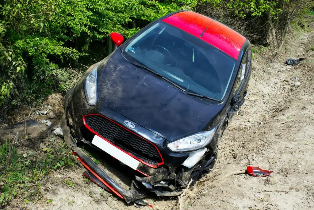 A damaged black and red car rests in a ditch surrounded by greenery. The front of the vehicle is severely dented, with debris visible around. The car appears to have skidded off the road and its license plate is blank.