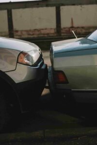 Close-up of a minor car collision with the front bumper of a silver SUV touching the rear bumper of a light green sedan. The vehicles appear to be parked on a quiet, dimly lit street with a concrete barrier in the background, raising concerns about potential insurance claims.