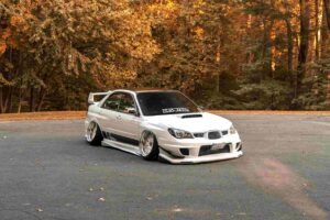 A modified white car is parked on an asphalt surface with a backdrop of autumn trees. The car features a body kit, a large rear wing, custom wheels, and a decal on the windshield. Remember to secure the right insurance for such bespoke modifications amid the golden leaves and fall setting.