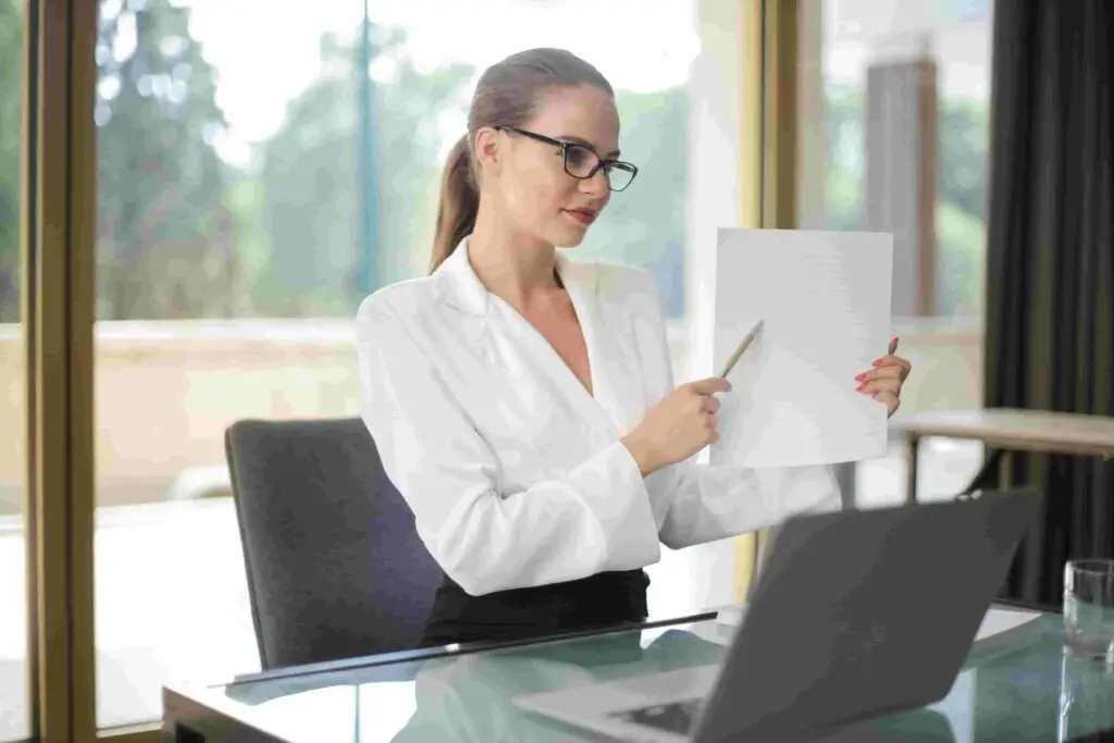 A woman wearing a white blouse and glasses is sitting at a desk, holding up a piece of paper in one hand and pointing to it with a pencil in the other. She is looking at a laptop screen, which displays an insurance document, while natural light streams through large windows behind her.