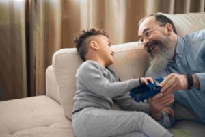 A young boy with curly hair and a light gray outfit plays with a toy car while sitting on a beige couch. An older man with a beard and glasses, wearing a light blue shirt, smiles and interacts with the boy. Beige curtains are in the background, making for a heartwarming scene reminiscent of an insurance commercial.