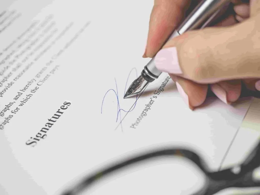 Close-up of a hand holding a fountain pen, signing an insurance document. The document has the heading "Signatures" and appears to be an agreement or contract. A pair of eyeglasses is partially visible in the lower part of the image.